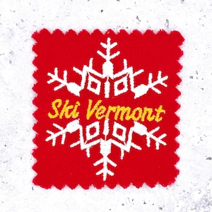 May include: A red fabric patch with a white snowflake design and the words "Ski Vermont" in yellow. The patch has a zig-zag edge.