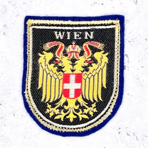 May include: A rectangular patch with a blue border and a black center. The patch features the word "WIEN" at the top and a detailed emblem with a red and white cross, a crown, and a golden eagle design.