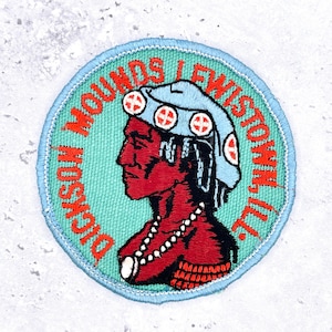 May include: A circular embroidered patch featuring a profile of a person with red skin wearing a light blue headdress with white and red circular accents. The text Dickson Mounds Lewistown, Ill. is stitched in red around the light blue background.
