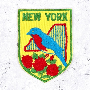 May include: A shield-shaped embroidered patch with a yellow background and a green border. The text NEW YORK is stitched in green at the top. It features a blue and red bird, a blue and yellow striped shape, and three red roses with green leaves.