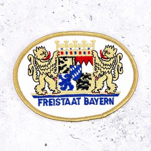 May include: Oval patch with a gold border featuring the Bavarian coat of arms. Two gold lions flank a shield with a crown, red, blue, and black elements. The text "FREISTAAT BAYERN" is in blue.