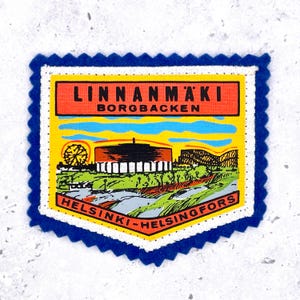 May include: A rectangular fabric patch with a blue scalloped border featuring a graphic illustration of Linnanmäki amusement park. The design includes a Ferris wheel, a building, and text reading LINNANMÄKI BORGBACKEN and HELSINKI-HELSINGFORS.