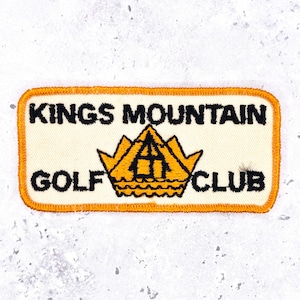 May include: A rectangular embroidered patch with an off-white background and a bright orange border. The text KINGS MOUNTAIN GOLF CLUB is stitched in black thread. A central yellow and black mountain graphic sits between the words.