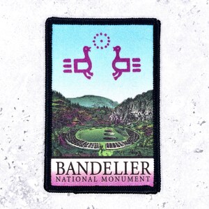 May include: A rectangular embroidered patch with a black border. The top section features two purple stylized birds facing each other against a light blue sky. The bottom depicts a green valley landscape with the text BANDELIER NATIONAL MONUMENT.