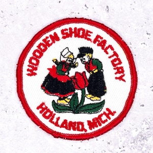 May include: Embroidered patch with a red border and the text "WOODEN SHOE FACTORY HOLLAND, MICH." in red. The patch features two cartoon figures in Dutch attire, a red tulip, and the words "Wooden Shoe Factory".