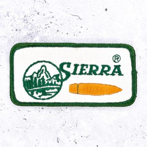 May include: A rectangular embroidered patch with a white background and a dark green border. It features a green circular mountain landscape logo, the word SIERRA in green text, and a horizontal orange bullet graphic with a registered trademark symbol.