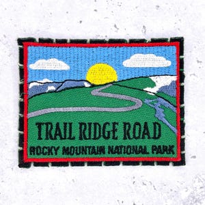 May include: Embroidered patch featuring a scenic landscape with a road winding through green hills under a blue sky with clouds and a yellow sun. The text "TRAIL RIDGE ROAD ROCKY MOUNTAIN NATIONAL PARK" is displayed.