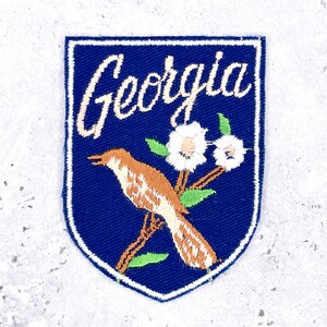 May include: A shield-shaped embroidered patch with a dark blue background and a white border. The text Georgia is stitched in white cursive at the top. Below, a brown bird sits on a branch with two white flowers and green leaves.
