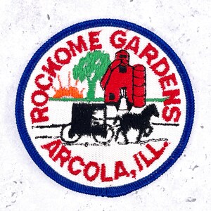 Op de afbeelding: Een cirkelvormige patch met een blauwe rand en een witte achtergrond. De patch heeft de tekst "ROCKHOME GARDENS ARCOLA, ILL." in het rood. Een rode schuur, groene boom en paard en wagen staan in het midden.