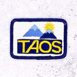 May include: Embroidered patch featuring the word "TAOS" in yellow block letters over a blue background. The patch includes a mountain range and sun graphic in blue and yellow, with a white background and a blue border.