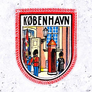 May include: A shield-shaped embroidered patch with a red border featuring the text KØBENHAVN at the top. The illustration depicts two guards in red uniforms and tall black hats standing near a red sentry box and a building with a blue window.