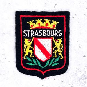 May include: Embroidered patch featuring the city name "STRASBOURG" in white, above a shield with a red diagonal stripe. The shield is flanked by yellow lions and topped with a yellow and red crown, all on a black background.