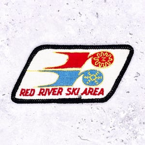 Pode incluir: Patch bordado com o texto "RED RIVER SKI AREA". O patch apresenta um fundo branco com formas estilizadas vermelhas e azuis, e um desenho de sol e floco de neve amarelo. O patch tem uma borda preta.