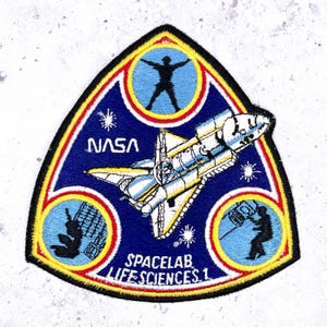 May include: A triangular patch featuring a space shuttle and the text "NASA" and "SPACELAB LIFE SCIENCES 1". The patch has a blue background with yellow and red borders, and includes illustrations of astronauts and scientific equipment.