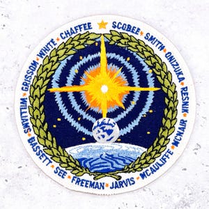 May include: Embroidered patch with a circular design featuring a starburst, a globe, and names around the edge. The patch has a white border, a blue background, and gold, white, and green accents. The names include Chaffee, Scobee, and Smith.