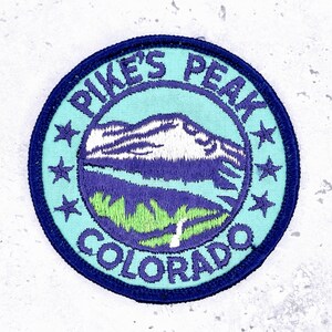 May include: Round embroidered patch featuring a mountain scene with the text "PIKE'S PEAK COLORADO". The patch has a teal background with a dark blue border and stars. The mountain is white and purple.