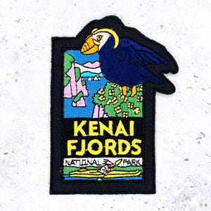 May include: Embroidered patch featuring a colorful illustration of a puffin perched above a scenic landscape. The patch includes the text "KENAI FJORDS NATIONAL PARK" in yellow and white lettering, set against a black border.