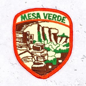 May include: Embroidered patch with the text "MESA VERDE" in green on a cream background. The patch features a brown and green landscape design with an orange border.