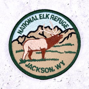 Puede incluir: Parche bordado circular con borde verde, que representa un uapití en un entorno natural. El parche incluye el texto "NATIONAL ELK REFUGE JACKSON WY". El uapití es marrón y blanco con cuernos, sobre un fondo de montañas y cielo azul claro.