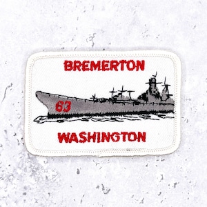 Peut inclure: Un écusson brodé rectangulaire à fond blanc avec une bordure cousue. Il présente un graphique de navire de guerre gris avec le numéro 63 en rouge sur la coque. Le texte BREMERTON est brodé en rouge au-dessus et WASHINGTON en rouge en dessous.