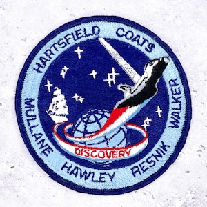 May include: Embroidered patch with a blue and white design. The patch features the text "Hartsfield Coats Walker Resnik Hawley Mullane" around the edge. A stylized image of a ship, a globe, and a space shuttle are in the center. The word "Discovery" is also present.