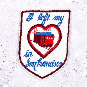 May include: Embroidered patch with a white background and a shield shape. The patch features a red heart with a red cable car inside. The text "I left my in San Francisco" is embroidered in blue.