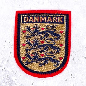 以下が含まれることがあります： 紺色の背景に金色の文字で「DANMARK」と刺繍された盾形のワッペン。中央には、金色のメタリックなフィールドに縦に並んだ3頭の紺色のライオンが描かれており、その周囲を9つの小さな赤いハートが囲んでいます。