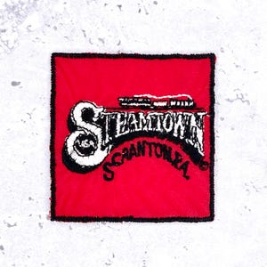 May include: A square red embroidered patch with a black stitched border. The design features the white text Steamtown in a stylized font, with the word Scranton, PA in black lettering underneath. A small white train graphic sits above the text.