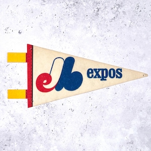 May include: A vintage Expos pennant in a triangular shape. The pennant is cream-colored with a red border and yellow tabs. The Expos logo is in red, white, and blue, with the word "expos" in blue.