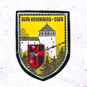May include: A shield-shaped patch with the text "BURG HOHENBERG/EGER" at the top. The patch features a yellow background with a white castle, a red and black shield, and green trees. The patch is bordered by a green edge.