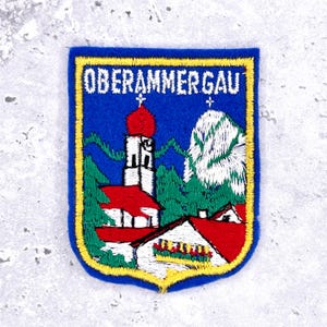 May include: Embroidered patch with the text "OBERAMMERGAU" in white on a blue background. The patch features a church with a red roof and a mountain scene, framed by a yellow border.