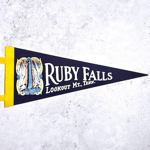 May include: A vintage pennant in navy blue with the words "RUBY FALLS LOOKOUT MT., TENN." in white. The pennant features a colorful illustration of Ruby Falls. The pennant has a yellow border and hanging tabs.