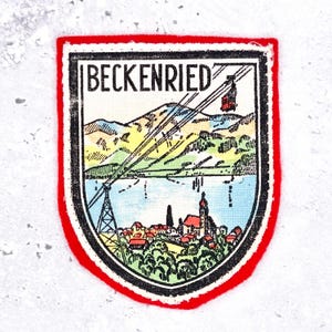 May include: A shield-shaped embroidered patch with a red border featuring a printed illustration of the town of Beckenried, Switzerland. The design includes a cable car, mountains, a lake, and a church, with the text BECKENRIED at the top.