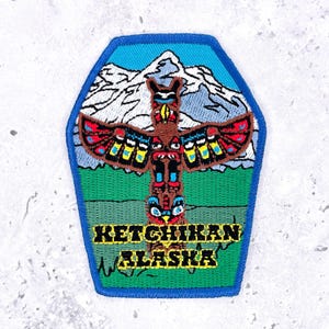 May include: A shield-shaped embroidered patch featuring a brown totem pole with red, yellow, and blue details centered against a backdrop of white snow-capped mountains and a green landscape. The text KETCHIKAN ALASKA is stitched in yellow.