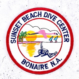 Peut inclure: Un écusson brodé circulaire à bordure rouge portant le texte SUNSET BEACH DIVE CENTER et BONAIRE N.A. en bleu foncé. Le motif représente un soleil jaune, un flamant rose, des palmiers, deux voiliers et un plongeur sous-marin en bleu.