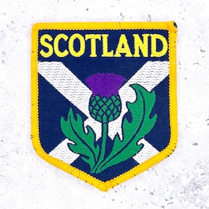 May include: Embroidered patch featuring the word "SCOTLAND" in yellow, above a white saltire on a navy blue background. A purple thistle with green leaves is centered on the patch, which has a yellow border.