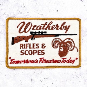 May include: A rectangular embroidered patch with a white background and a gold border. It features a brown rifle with a scope, a brown ram head, and the text Weatherby RIFLES & SCOPES and Tomorrow's Firearms Today in brown and red lettering.