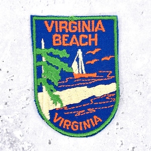 May include: A shield-shaped embroidered patch with a blue background and a green border. It features orange text reading VIRGINIA BEACH and VIRGINIA, a green tree, a white sailboat on orange water, and orange birds against the blue sky.
