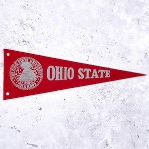 May include: A red felt pennant with the words "OHIO STATE" in silver lettering. The left side features a circular emblem with text including "THE OHIO STATE UNIVERSITY" and "COLUMBUS 1870". The pennant has two small holes at the top.