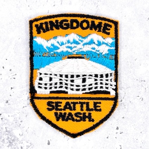May include: A shield-shaped embroidered patch featuring the text KINGDOME at the top and SEATTLE WASH. at the bottom in black thread. The center depicts a white domed stadium against a backdrop of blue mountains and a light blue sky.