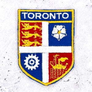 May include: A shield-shaped embroidered patch featuring the word TORONTO in white letters on a blue background. The crest is divided into four quadrants with a white cross, displaying three yellow lions, a white rose, a white gear, and a yellow ship.