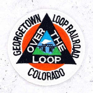 Puede incluir: Un parche bordado circular con un triángulo negro sobre un círculo naranja. El texto dice GEORGETOWN LOOP RAILROAD, OVER THE LOOP y COLORADO. Dentro del triángulo hay un cielo azul, una nube blanca, un viaducto negro y colinas verdes.