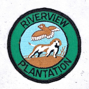 May include: Embroidered patch with the words "RIVERVIEW PLANTATION" in green and white. The patch features a brown bird in flight above a brown and white dog. The background is light blue and the border is black.