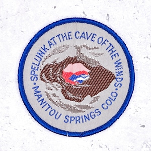 May include: A round patch with a blue border and the text "MANITOU SPRINGS COLO" and "SPELUNK AT THE CAVE OF THE WINDS" in blue. The center features a brown cave illustration with red and blue accents.