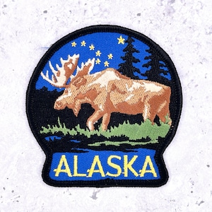 以下が含まれることがあります： 星空の下、緑の野原を歩く茶色のヘラジカを描いた刺繍パッチ。パッチには2本の濃い緑色の木と、青いバナーに黄色の文字で「ALASKA」の文字が含まれています。