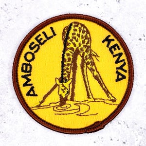 May include: Embroidered patch featuring a giraffe drinking water, with the words "AMBOSELI KENYA" in a circular design. The patch has a yellow background, a brown border, and the giraffe is brown with yellow spots.