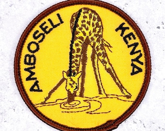 Op de afbeelding: Geborduurde patch met een giraffe die water drinkt, met de woorden "AMBOSELI KENYA" in een cirkelvormig ontwerp. De patch heeft een gele achtergrond, een bruine rand en de giraffe is bruin met gele vlekken.