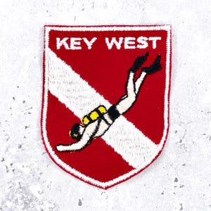Könnte beinhalten: Gestickter Aufnäher mit rotem Hintergrund und weißem diagonalen Streifen. Der Aufnäher zeigt den Text "KEY WEST" in Weiß und eine Illustration eines Tauchers. Der Taucher trägt einen schwarzen Neoprenanzug und gelbe Tauchausrüstung.