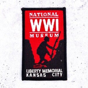 May include: A rectangular embroidered patch with a black border. The design features a red background with white text reading NATIONAL WWI MUSEUM. Below, a black silhouette of a soldier climbing a hill. At the bottom, white text reads LIBERTY MEMORIAL KANSAS CITY.