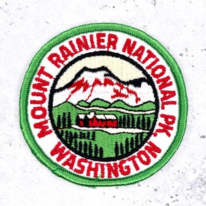 May include: Embroidered patch featuring Mount Rainier National Park in Washington. The design includes a mountain range with red and white peaks, green hills, and a red building. The text "Mount Rainier National Park Washington" is in red.
